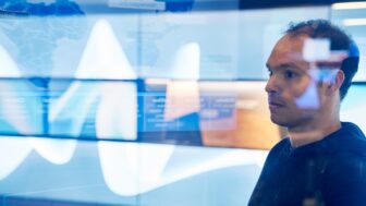 Man in sweater inside a secure room who is looking at data and a geographic area displayed on a large monitor which is behind glass walls with reflections.