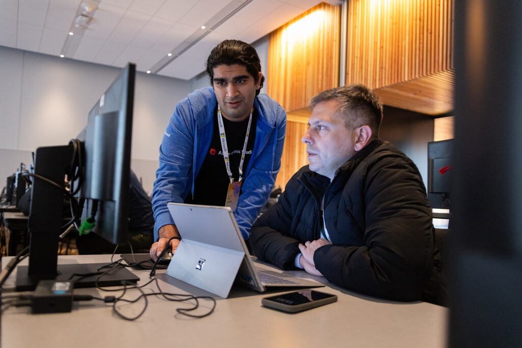 Two people sit at a conference looking at a computer screen and a tablet. 