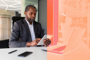 A man sits in front of a laptop holding a tablet.