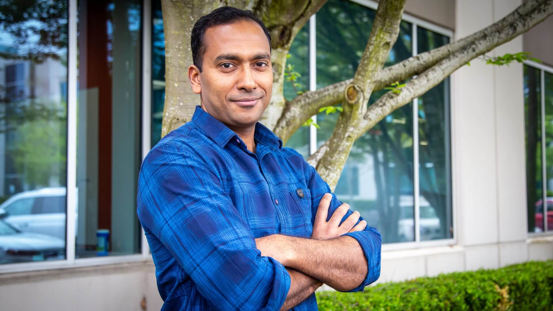 Garg stands in front of a large tree and building with his arms crossed, and he smiles at the camera.