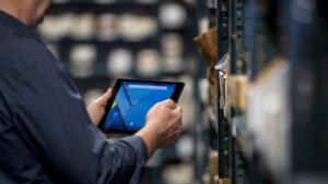 Male employee holding a tablet with both hands. He is standing in front of a metal warehouse rack filled with packages.