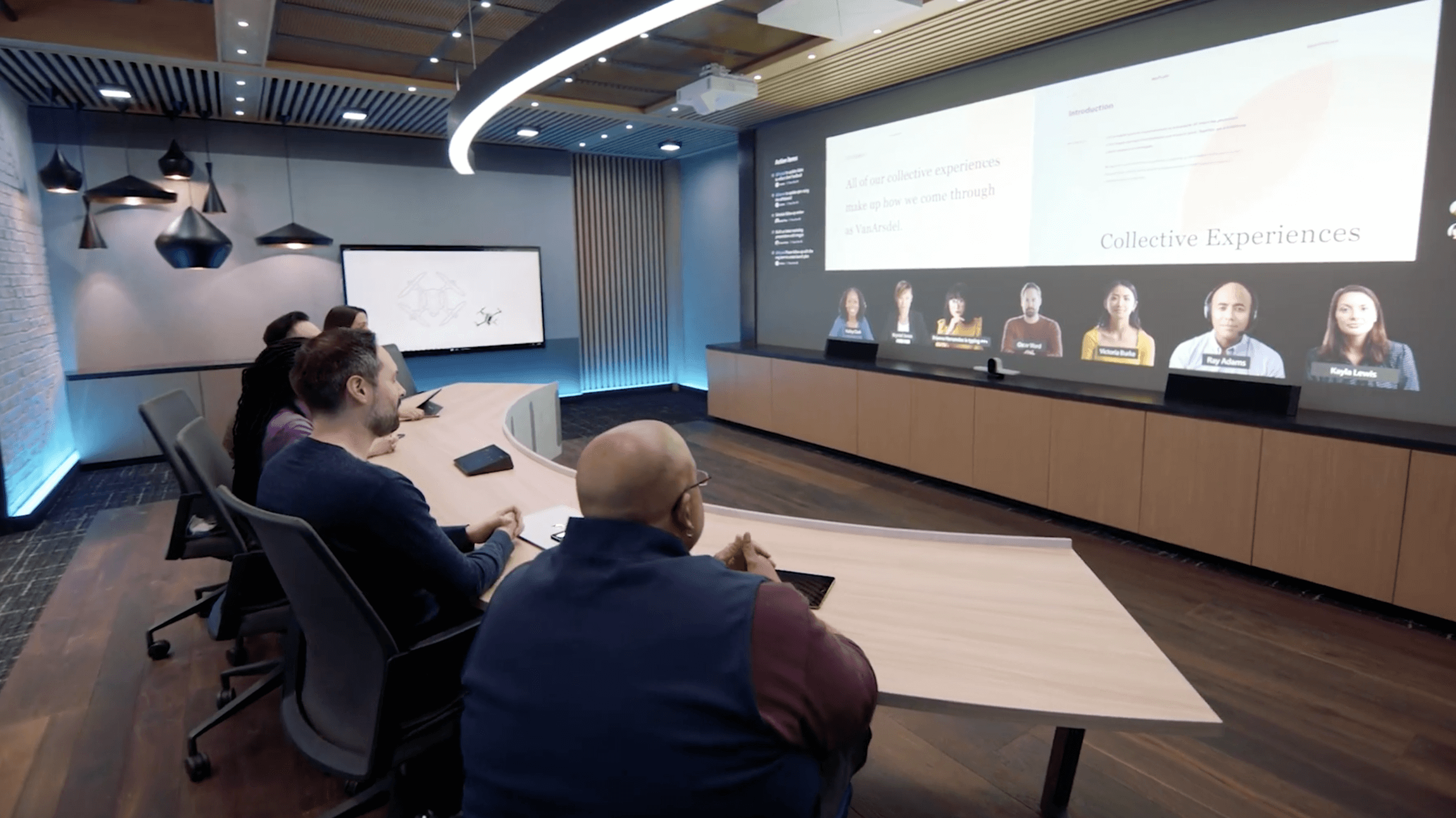 Microsoft employees are shown at a curved table facing a screen as people in the room and joining remotely meet.