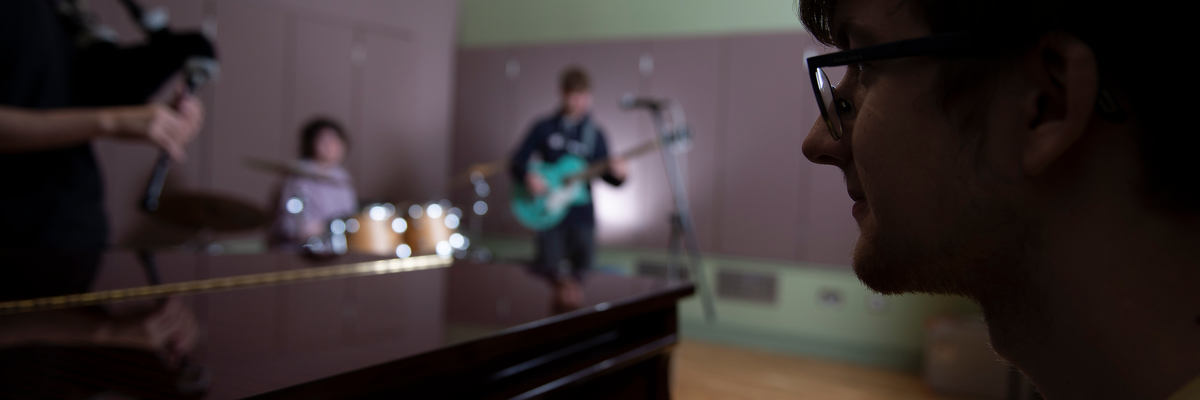 Male student at a piano with a band playing in the background