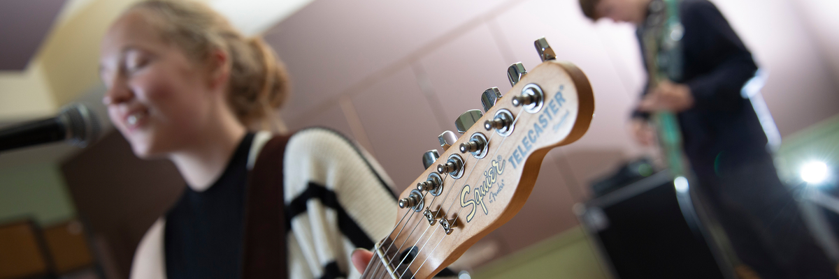 Female student standing in front of a microphone while playing a guitar