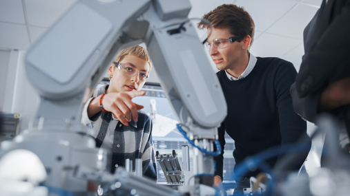 A man and a woman talk while the woman is pointing at a robotic arm