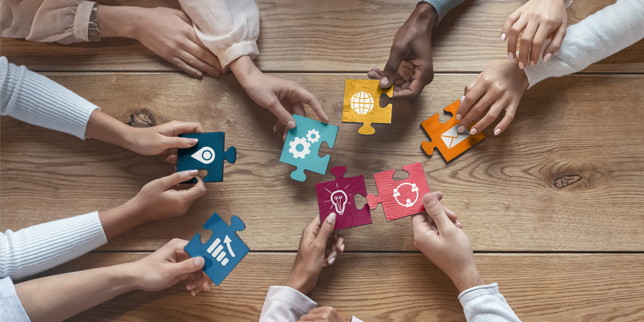 Several hands holding puzzle pieces with various work-related icons, reaching toward each other across a wooden table to connect the pieces into one image. 