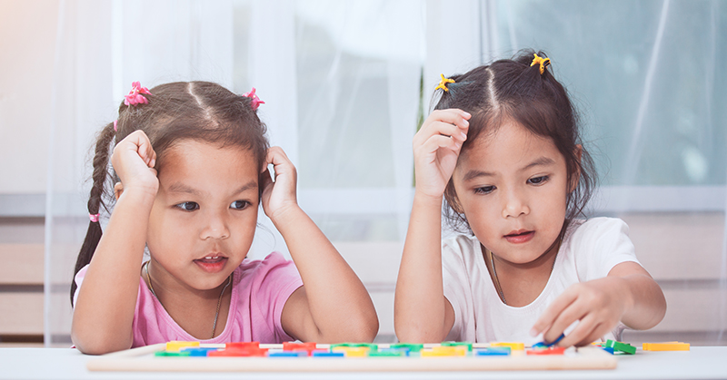 Two children seated at table. One inserts a piece to a puzzle while the other watches.