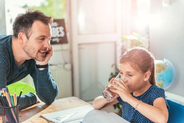 Padre mirando a su hija tomar un vaso de agua mientras hace las tareas escolares.