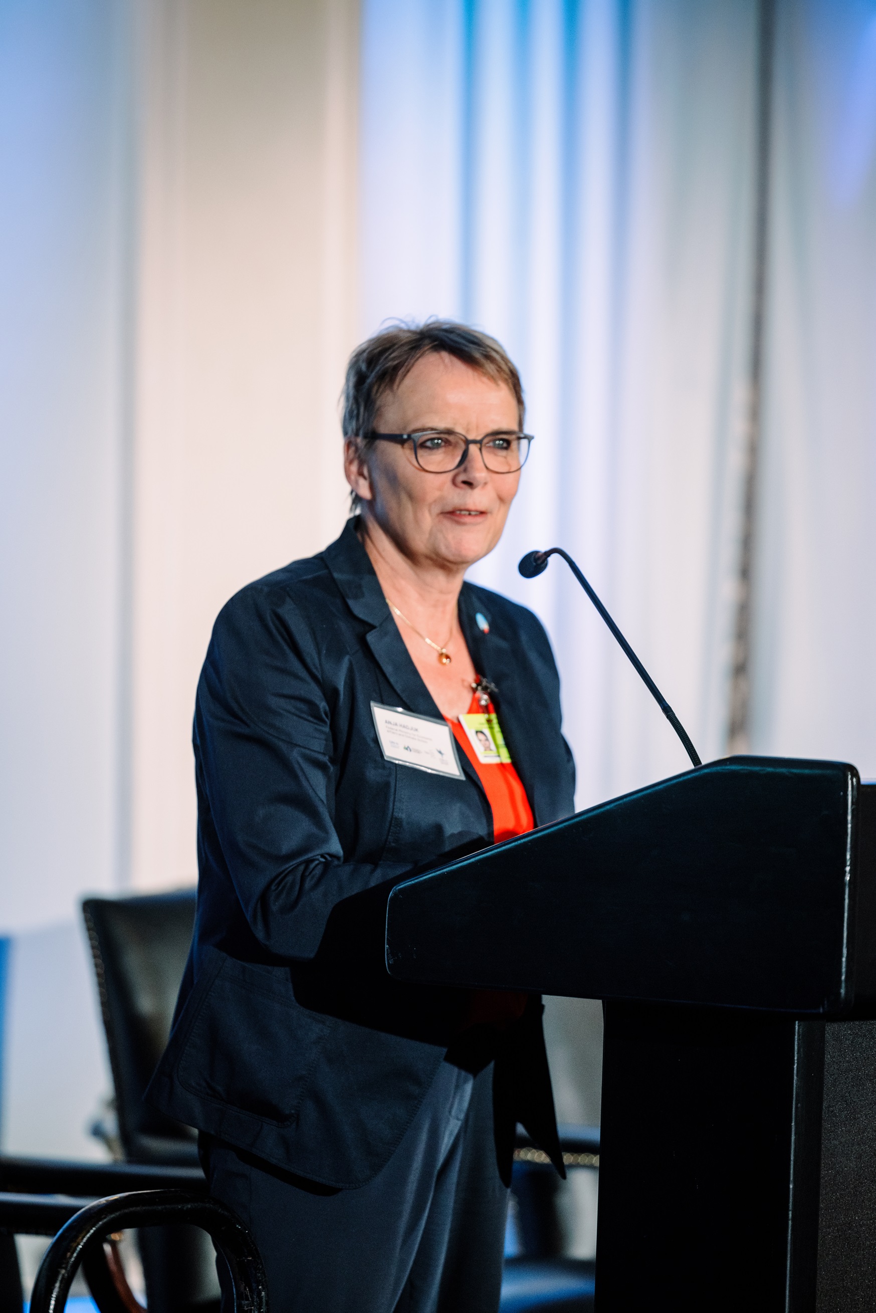 Middle-aged woman with dark readin glasses and short gray hair, wearing a dark blue blazer and a red shirt, speaks in a podium