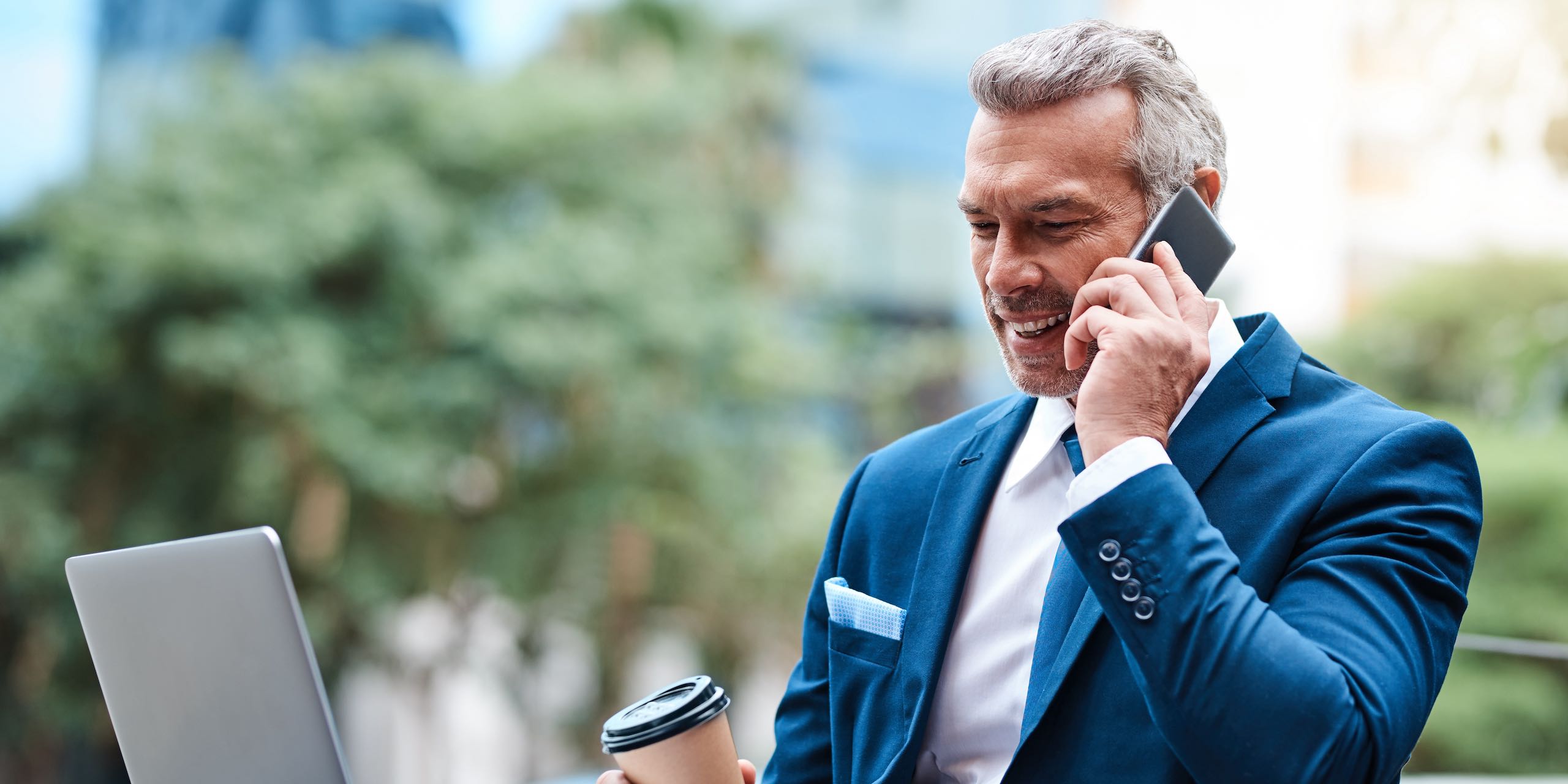 Shot of a handsome mature businessman in corporate attire on a call and using a laptop outside during the day