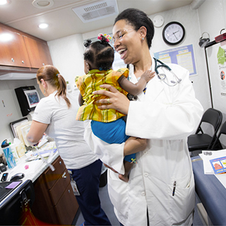 In an examination room, a smiing medical school student with a stethoscope around her neck holds a baby