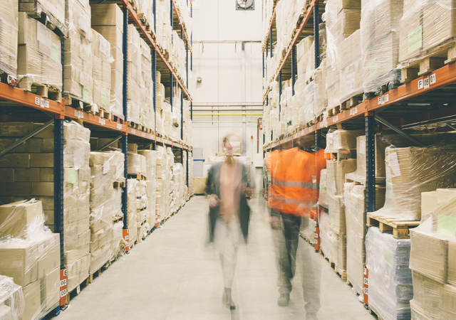 Two people walking through a warehouse of packages on shelves