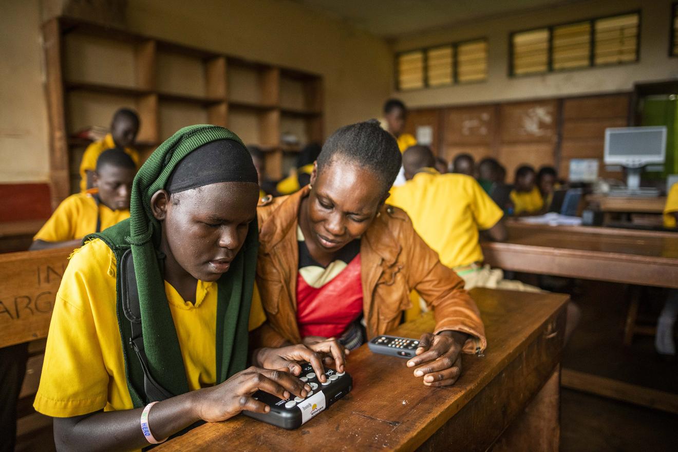 Une professeure assise à côté de l'une de ses élèves pour l'aider à utiliser un lecteur optique de braille