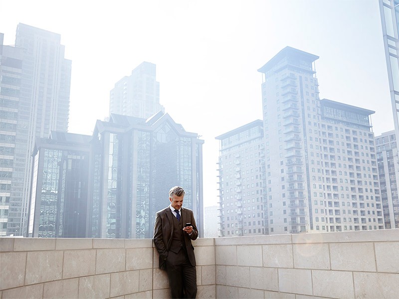 Businessman in a suit checking phone on a rooftop terrace, surrounded by urban skyline of tall modern buildings 