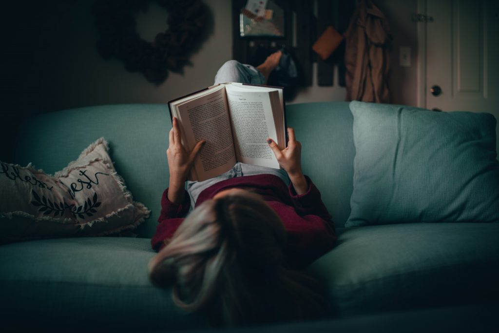 Woman is laying upside down in sofa and reads a book.