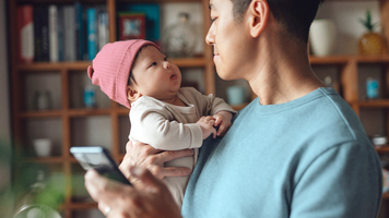 Asian father checking emails on smartphone while playing with her baby. Parent working from home. Remote working concept.