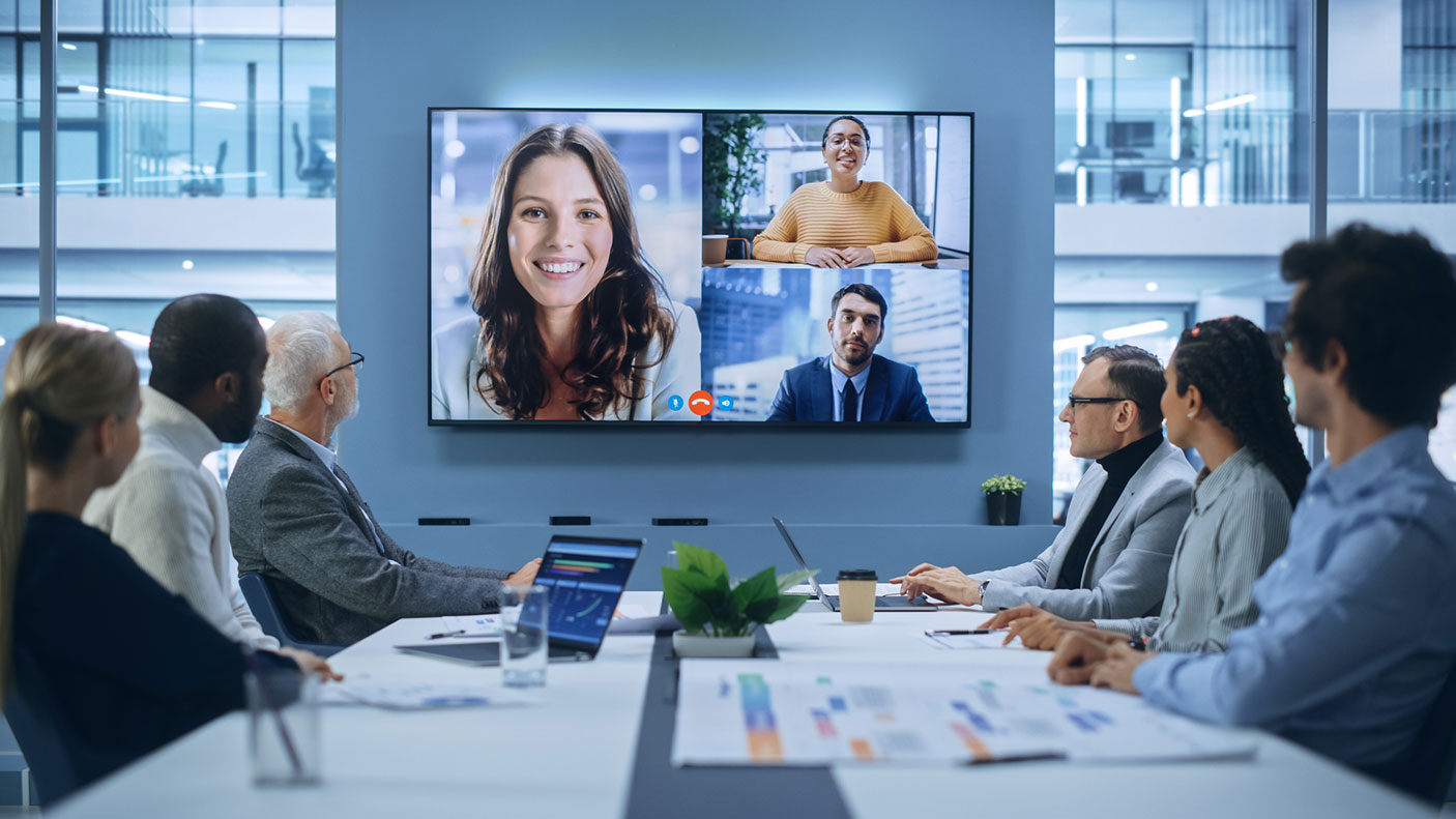 A group of people in a meeting room are looking at the TV screen which has three meeting attendees online.